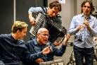 Een oudere heer maakt kunst door op een trommel te spelen en te zingen in een microfoon. Deze wordt hem voorgehouden door een jongere heer. Op de achtergrond bespelen meer mensen een muziekinstrument