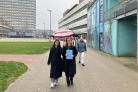 Deelnemers aan het werkbezoek lopen lachend in de regen onder een paraplu op het terrein van de VU.