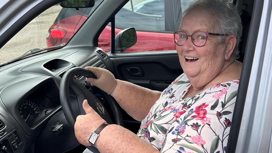 Lachende oudere dame achter stuur in auto, ze steekt haar duim op