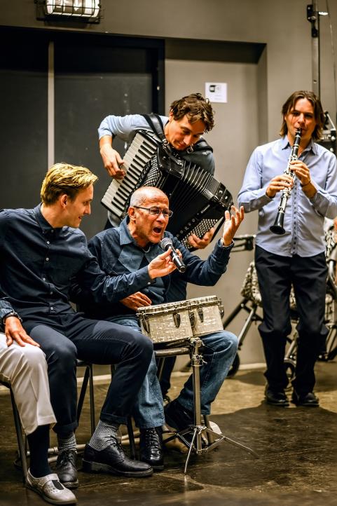 Een oudere heer maakt kunst door op een trommel te spelen en te zingen in een microfoon. Deze wordt hem voorgehouden door een jongere heer. Op de achtergrond bespelen meer mensen een muziekinstrument