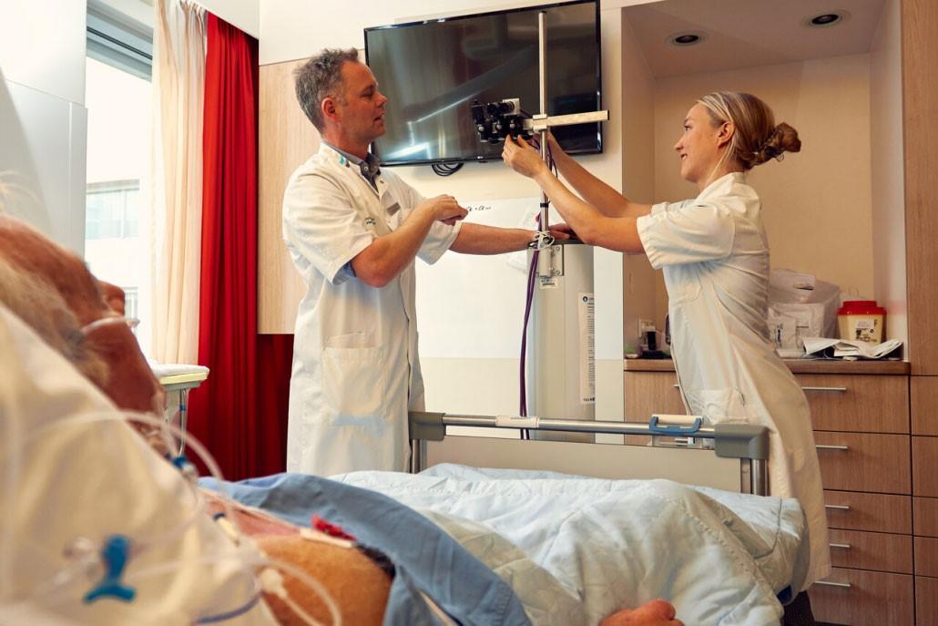 Twee verpleegkundigen stellen videocamera in aan voeteneind van bed waarin een patiënt ligt