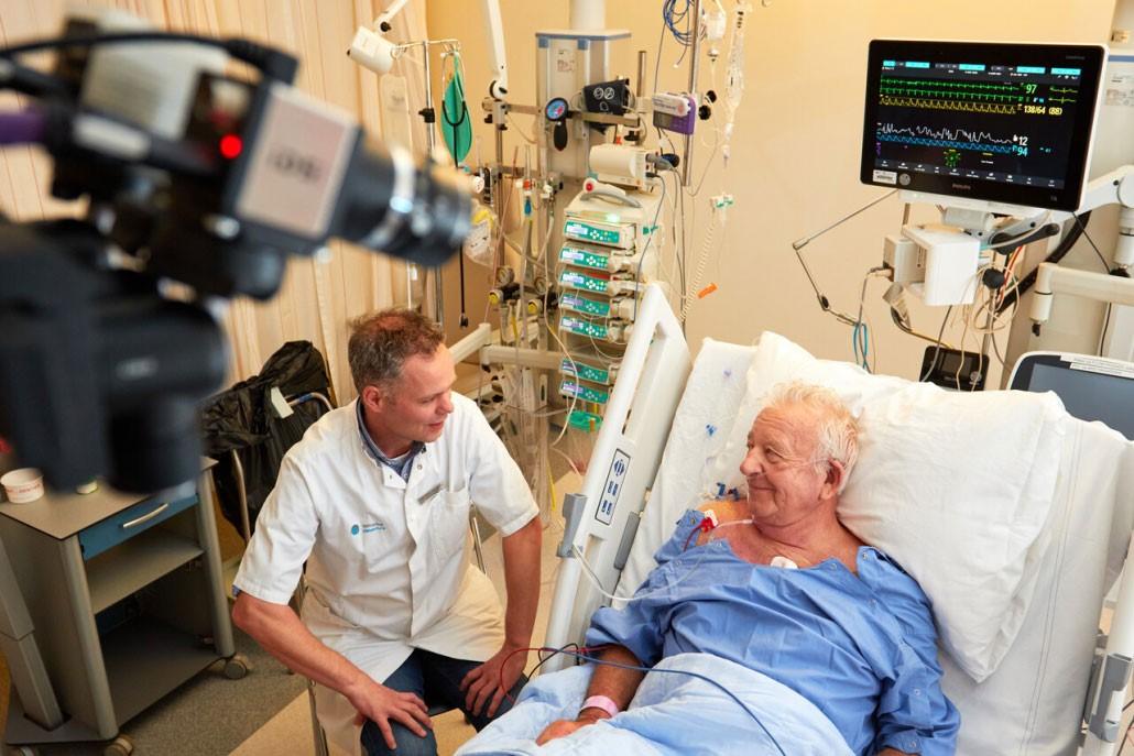 Verpleegkundige praat met patiënt die in bed ligt, op de voorgrond een videocamera