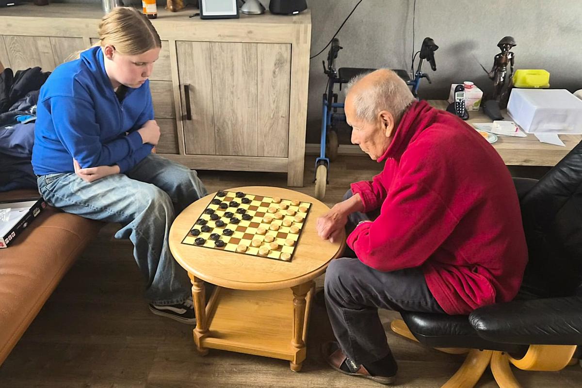 Een jongere dame en oudere heer spelen in huiselijke omgeving een potje dammen.