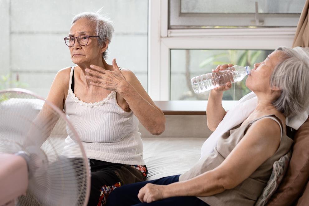Twee vrouwen zitten in een huis voor de ventilator. Het is duidelijk warm. Een vrouw wappert met haar hand om af te koelen en de andere vrouw drinkt een glas water.