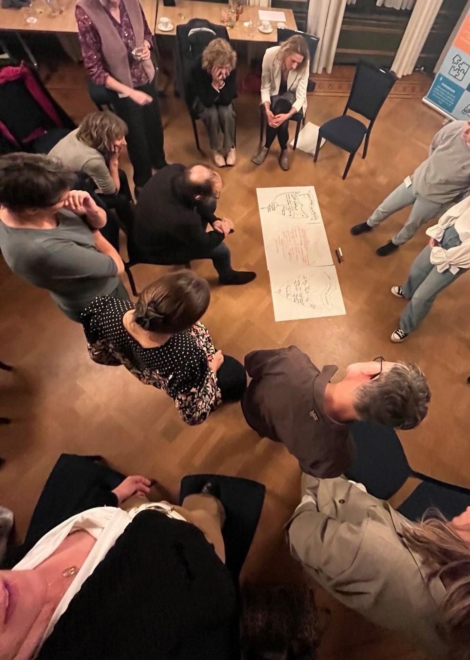 Mensen staan en zitten in een kring rond een groot vel papier op de grond voor een opdracht van de SVP-II training