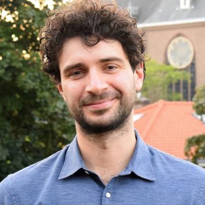 Man van gemiddelde leeftijd met middellang krullend, bruin haar met een blauwe blouse aan. Hij staat buiten in de stad